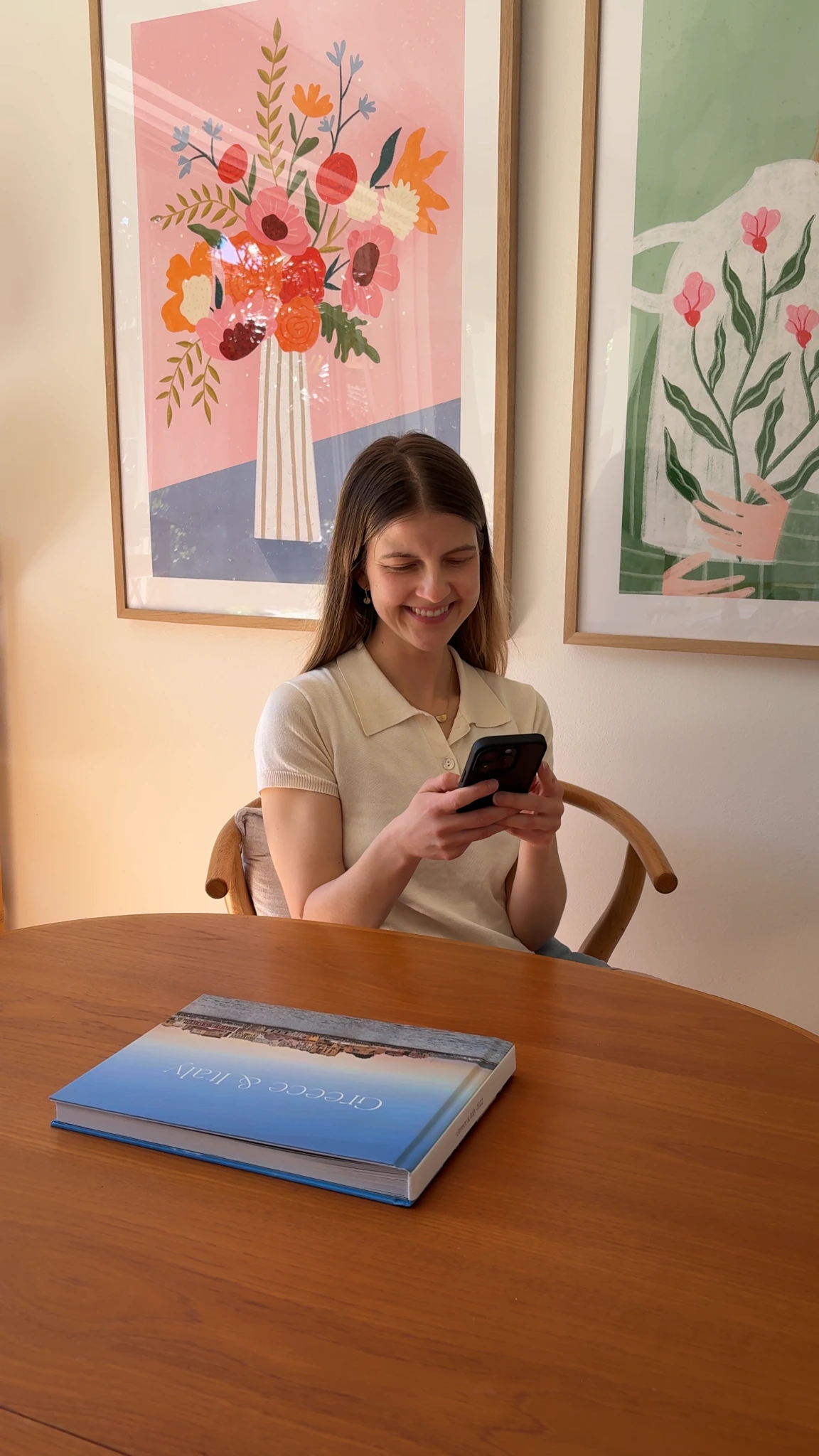 Alexis smiling at her phone at a dining table beneath two framed floral prints